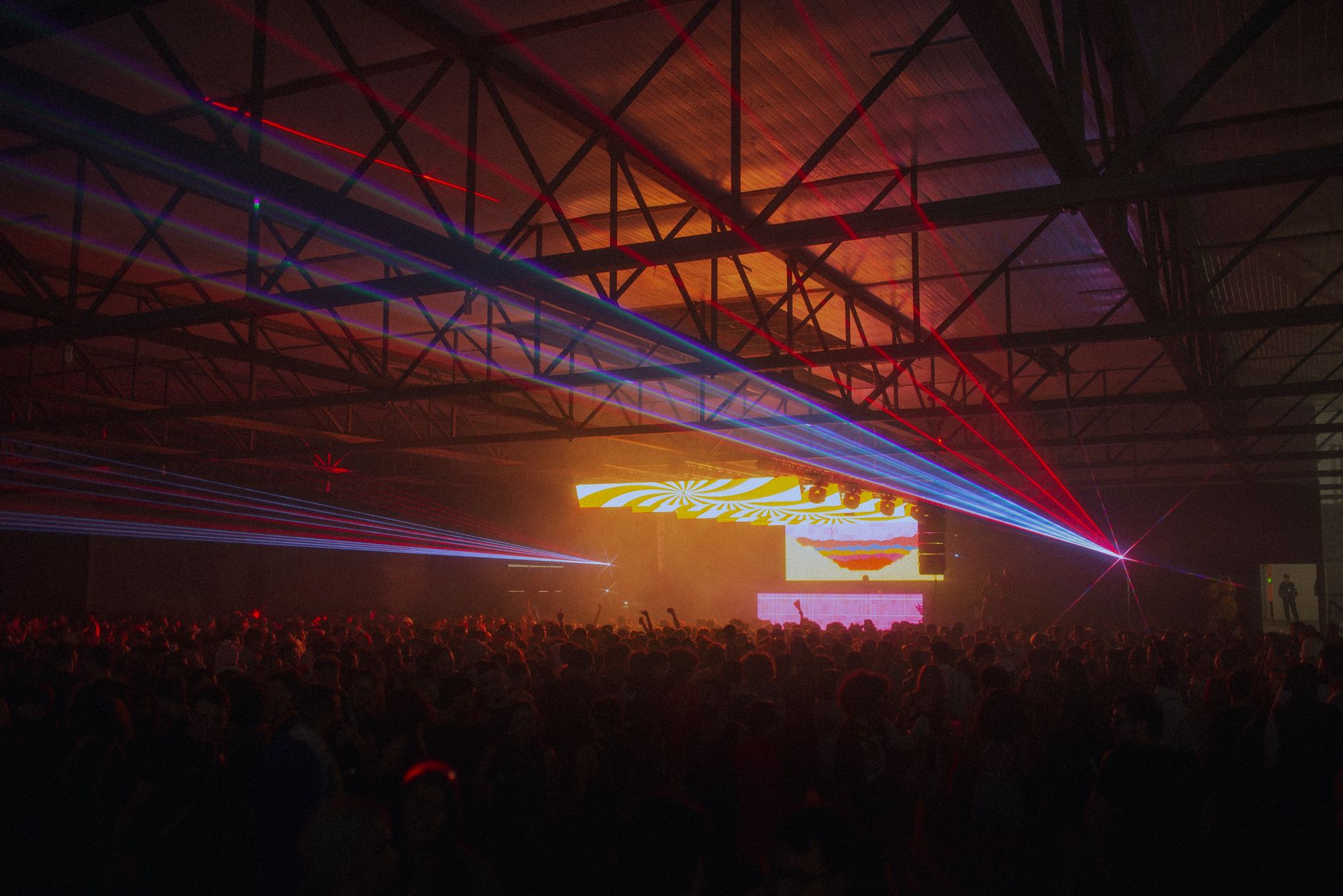 Laser beams crossing a warehouse arena above a dense crowd facing the stage