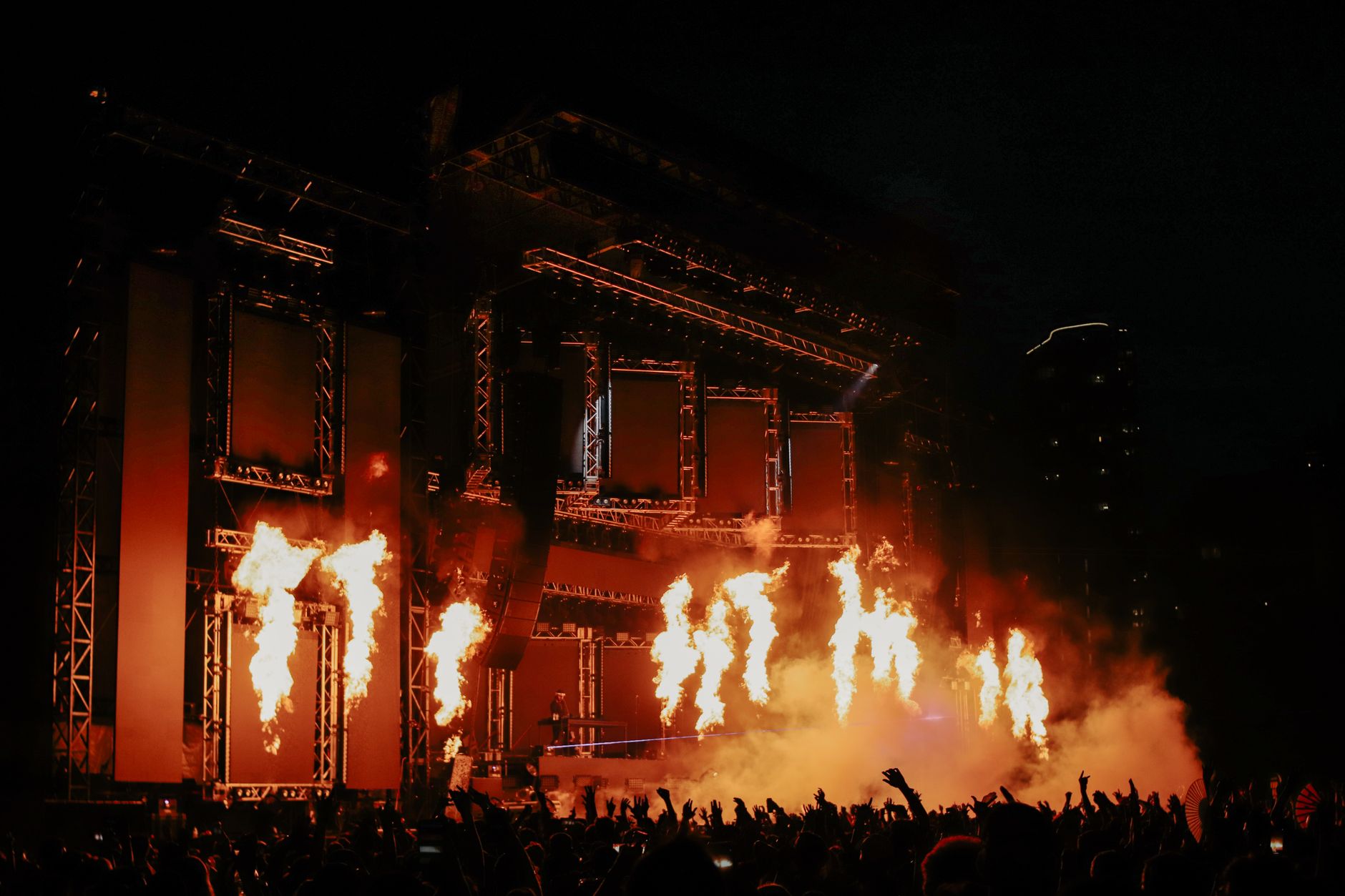 Six columns of stage pyrotechnics firing above a crowd with hands raised
